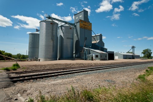 Grain silos and a processing facility under a clear blue sky, with railroad tracks in the foreground.