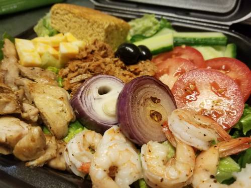 A colorful salad with grilled chicken, shrimp, tomatoes, onions, cheese, olives, cornbread, and lettuce.