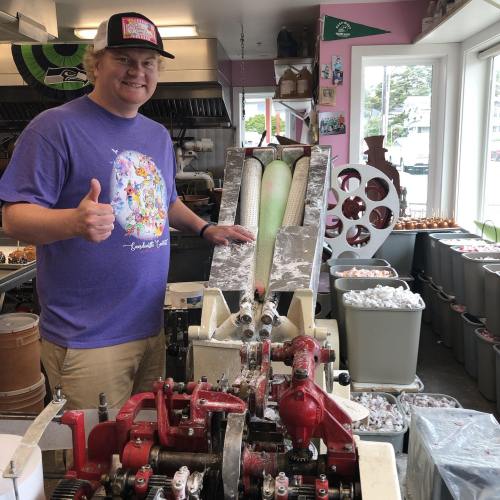 A smiling man in a purple shirt gives a thumbs up next to a vintage ice cream machine in a colorful shop.