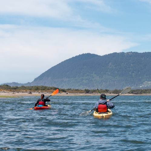 Two people kayaking on a calm waterway, with mountains and blue skies in the background.