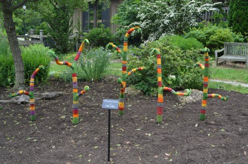 Colorful, knitted tree sculptures in a garden, with a sign labeled "Yarn." Lush greenery surrounds the installation.