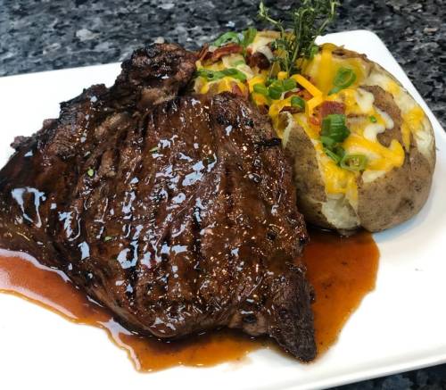 A juicy steak with a glossy sauce, served alongside a loaded baked potato topped with cheese, bacon, and green onions.