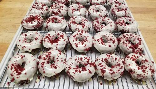 A tray of red velvet donuts topped with white icing and red crumbs, arranged neatly on a cooling rack.