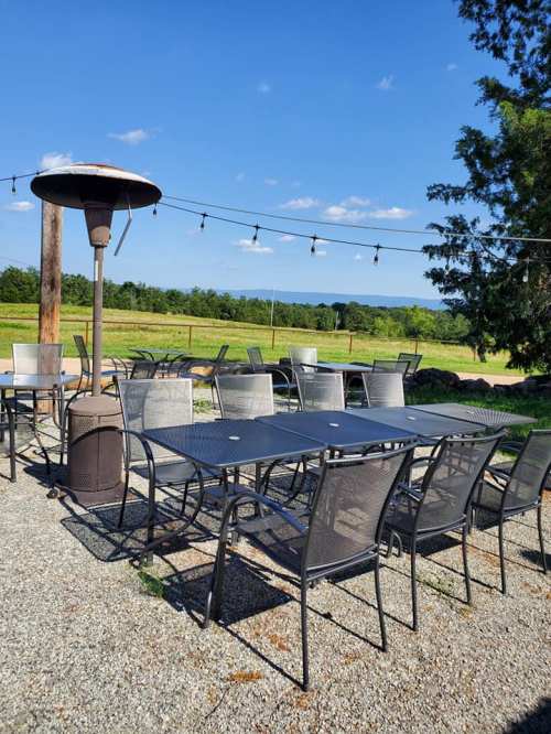 Outdoor dining area with metal tables and chairs, a patio heater, and string lights against a scenic green landscape.