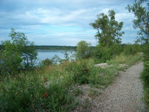 A serene lakeside scene with a gravel path, lush greenery, and a calm water surface under a cloudy sky.