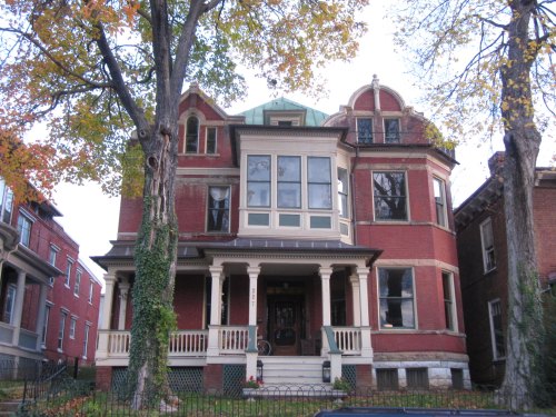 A large, historic red brick house with a green roof, featuring multiple windows and a front porch, surrounded by trees.