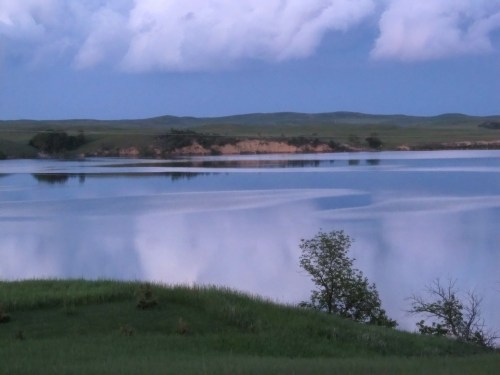 A serene lake reflects clouds and hills, surrounded by lush green grass and trees under a twilight sky.