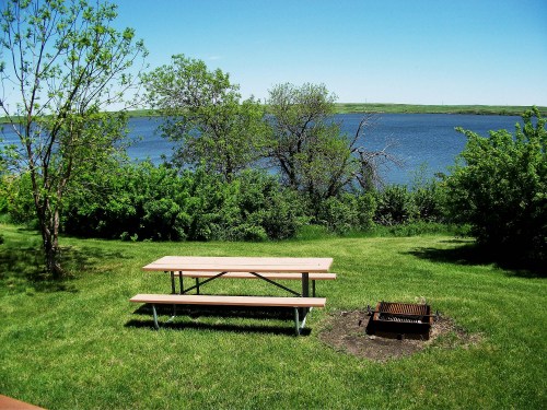 A serene lakeside view featuring a picnic table, grill, and lush greenery under a clear blue sky.