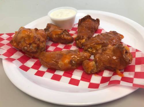 A plate of saucy chicken wings on a red and white checkered paper, accompanied by a small cup of ranch dressing.