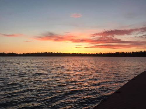 A serene sunset over a calm lake, with vibrant orange and pink hues reflecting on the water. Silhouetted trees in the background.