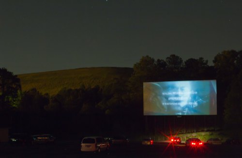 A drive-in movie screen displays text at night, with cars parked in front and a hillside in the background.
