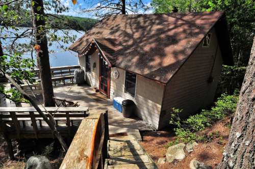 A cozy cabin by a lake, surrounded by trees, with a wooden deck and steps leading down to the water.