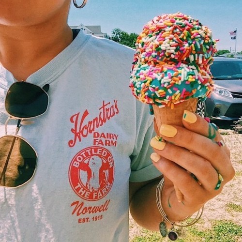 A person holds a colorful ice cream cone topped with sprinkles, wearing a light blue shirt and sunglasses.