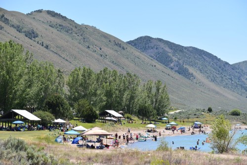 A sunny beach scene with people enjoying the water, surrounded by green trees and mountains in the background.