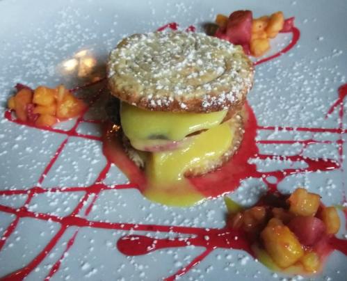 A dessert featuring two cookie-like layers filled with yellow cream, garnished with fruit and a red sauce drizzle.