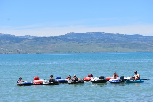 A group of people relaxes on inflatable rafts in a calm lake, surrounded by mountains and clear blue skies.