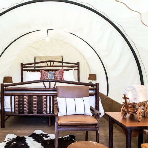 Cozy interior of a glamping tent featuring a bed, chair, and rustic decor with a wagon model on a table.