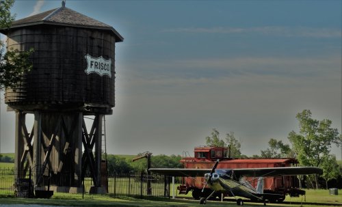 A vintage water tower labeled "Frisco" beside a classic airplane and an old train car in a rural setting.
