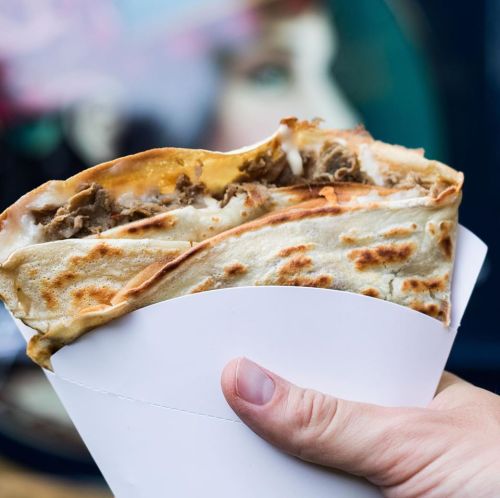 A hand holding a large, stuffed quesadilla with melted cheese and beef, set against a colorful background.