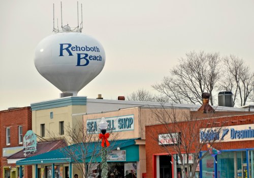 A water tower labeled "Rehoboth Beach" stands above colorful shops along a street in a coastal town.