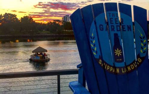 A blue chair with "The Beer Garden" logo overlooks a river at sunset, with a boat passing by in the background.