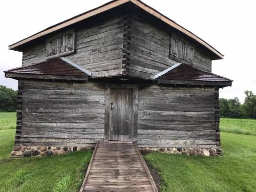 A rustic wooden cabin with a slanted roof, surrounded by green fields and a wooden walkway leading to the door.
