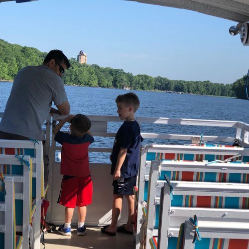 Two young boys and an adult stand on a boat, looking out at a calm lake surrounded by greenery.