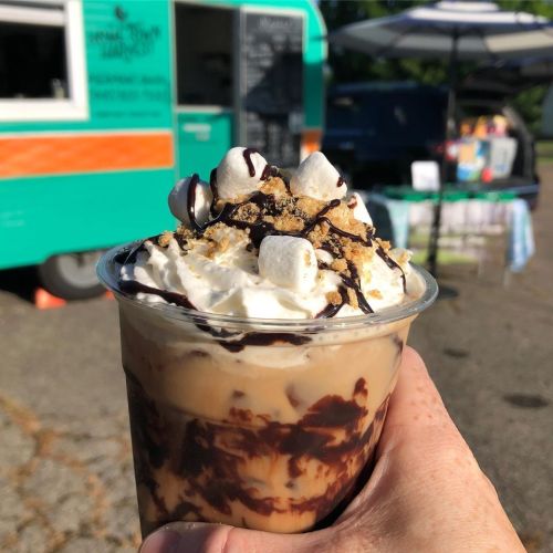 A hand holds a dessert cup filled with layers of chocolate, whipped cream, marshmallows, and graham cracker crumbs.