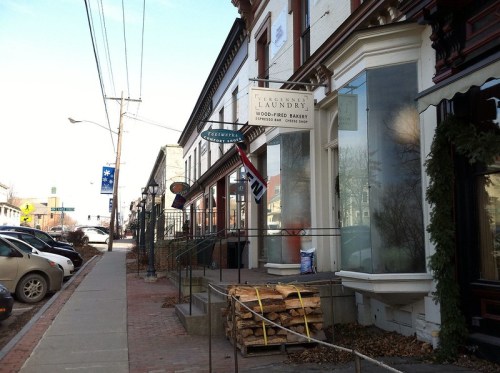 A quaint street scene featuring a bakery and laundry, with parked cars and stacked firewood in front.