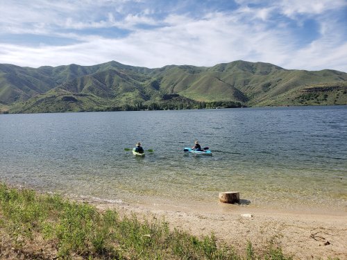 Two people kayaking on a calm lake surrounded by green mountains under a blue sky with scattered clouds.