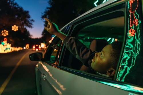 A child leans out of a car window, pointing excitedly at colorful holiday lights along a festive road.