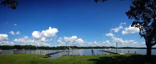 A serene lake scene with a clear blue sky, fluffy clouds, and a wooden dock surrounded by green grass and trees.