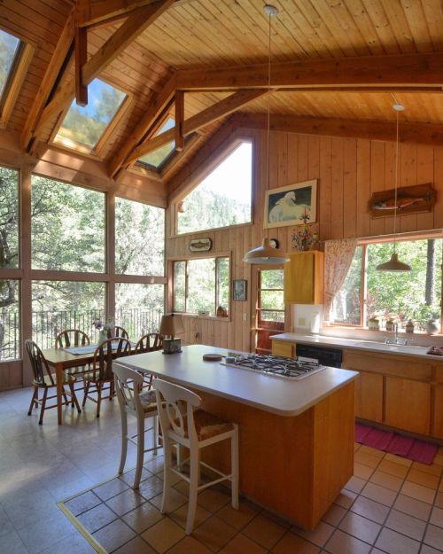Bright wooden kitchen with high ceilings, large windows, and a dining area, surrounded by trees. Cozy and inviting atmosphere.