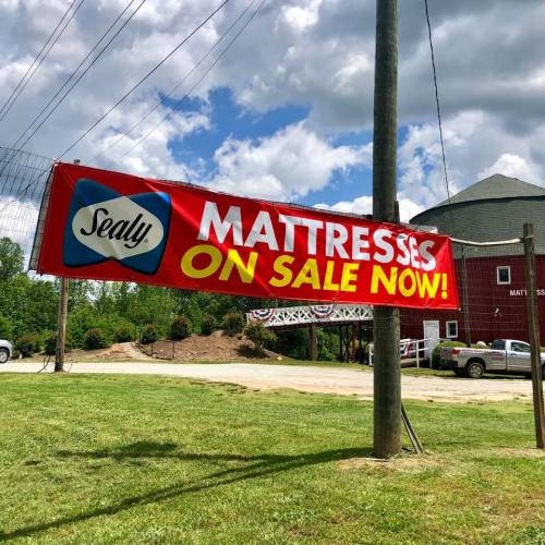 A large banner advertising Sealy mattresses on sale, displayed outside a building with a grassy area and cloudy sky.