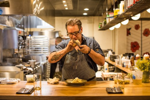 A chef in a kitchen prepares a dish, focusing intently as he garnishes a plate on a wooden countertop.
