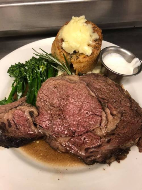 A plate featuring a juicy slice of prime rib, broccolini, a baked potato with sour cream, and herbs.