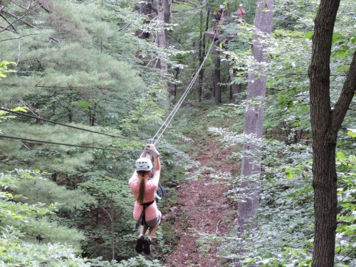 A person zip-lining through a forest, surrounded by trees and greenery, with another person visible in the background.