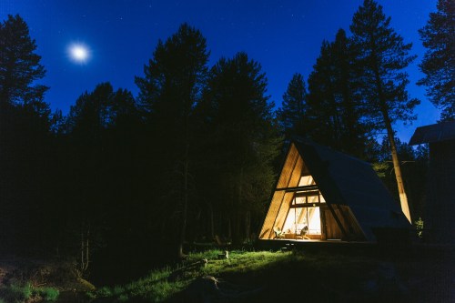 A cozy cabin illuminated at night, surrounded by tall trees and a starry sky, with a bright moon overhead.