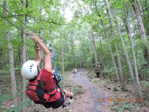 A person in a helmet zip lines through a forested area, with others visible on a path in the background.