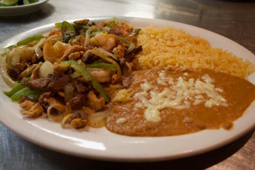 A plate of mixed grilled meats with peppers, served with rice and a side of cheese-topped refried beans.