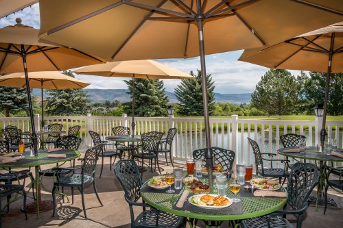 Outdoor dining area with green tables, umbrellas, and scenic views of trees and a pond. Plates of food and drinks on tables.