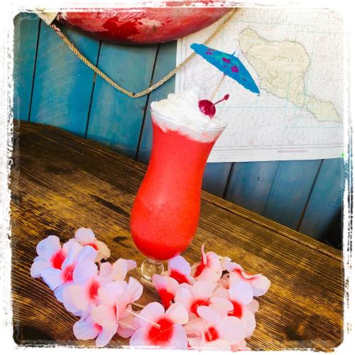 A vibrant pink cocktail with a cherry and umbrella, surrounded by pink flower petals on a wooden table.
