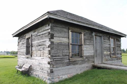 A rustic wooden cabin with a slanted roof, featuring barred windows and a small porch, set in a grassy landscape.