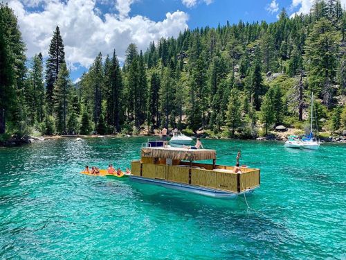 A floating wooden platform with people enjoying a sunny day on a clear blue lake surrounded by lush green trees.