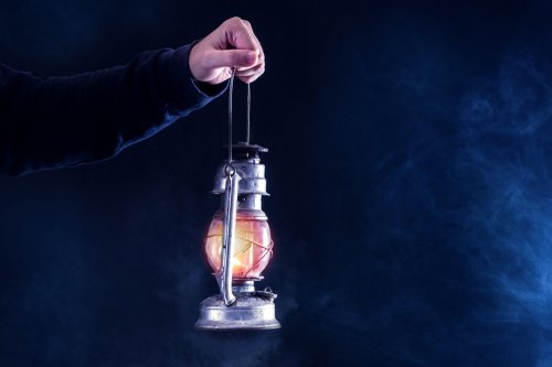 A hand holds an old-fashioned lantern, glowing warmly against a dark, smoky background.