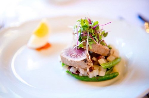 A gourmet dish featuring sliced tuna atop diced vegetables, garnished with microgreens and a lemon wedge.