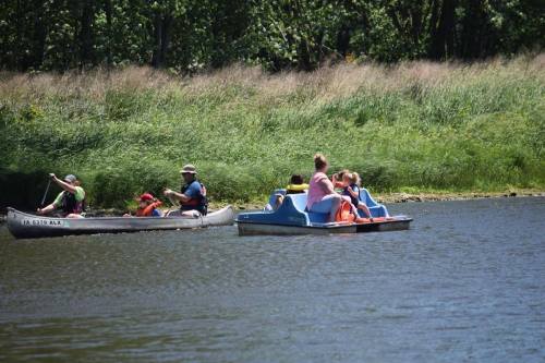 Two boats on a calm lake: one with paddlers and another with families enjoying a sunny day. Lush greenery in the background.