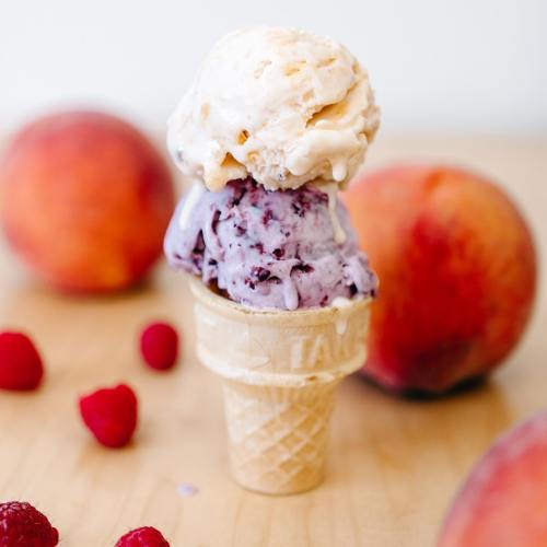 A cone with two scoops of ice cream, one peach and one berry, surrounded by fresh peaches and raspberries.