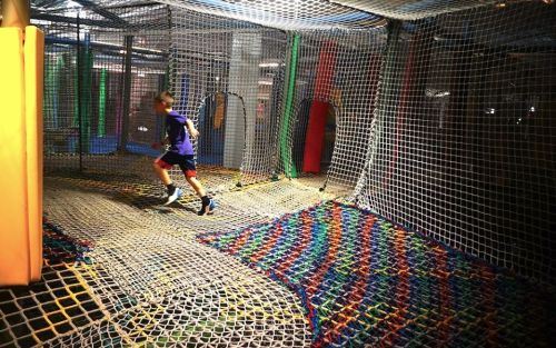 A child runs through a colorful netted play area with various tunnels and climbing structures.