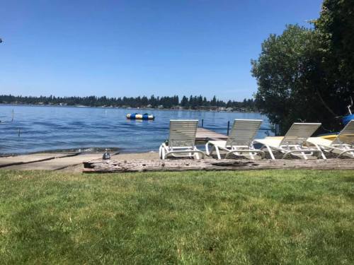 A serene lakeside view with lounge chairs on the grass, calm water, and trees in the background under a clear blue sky.
